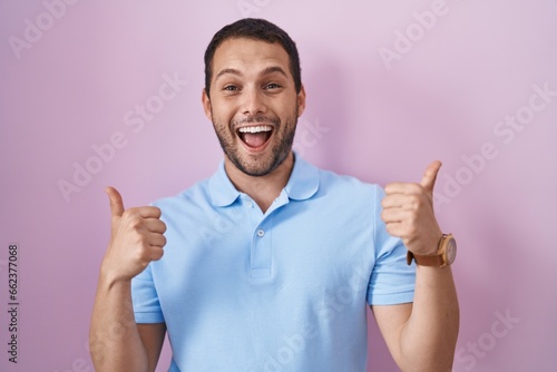 Hispanic man standing over pink background success sign doing positive gesture with hand, thumbs up smiling and happy. cheerful expression and winner gesture.