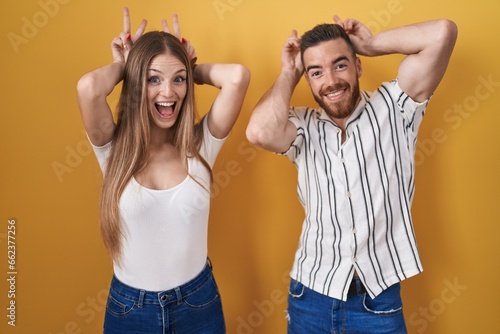 Young couple standing over yellow background posing funny and crazy with fingers on head as bunny ears, smiling cheerful