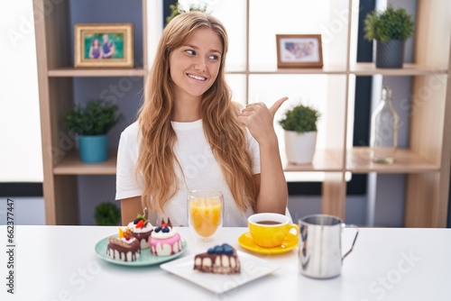 Young caucasian woman eating pastries t for breakfast smiling with happy face looking and pointing to the side with thumb up.
