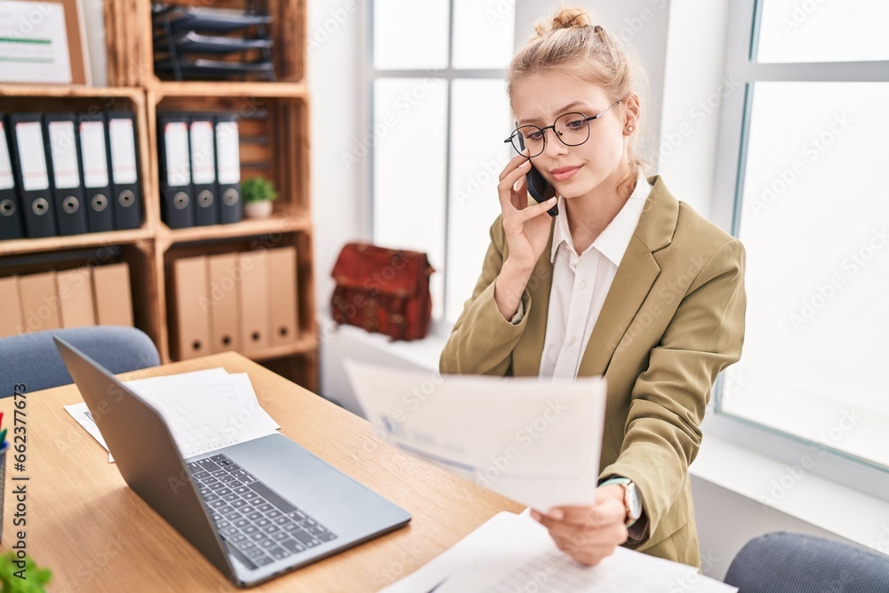 Young blonde woman business worker talking on smartphone reading document at office Young blonde woman business worker talking on smartphone reading document at office