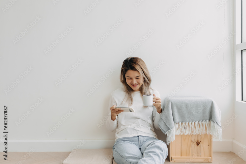 Asian Thai woman enjoy using and holding smartphone in apartment winter time. Happy moment smiling and chilling alone, sitting on floor, drink coffee. Asian Thai woman enjoy using and holding smartphone in apartment winter time. Happy moment smiling and chilling alone, sitting on floor, drink coffee.