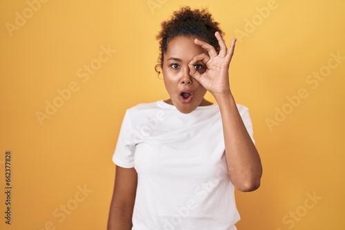 Young hispanic woman with curly hair standing over yellow background doing ok gesture shocked with surprised face  eye looking through fingers. unbelieving expression.