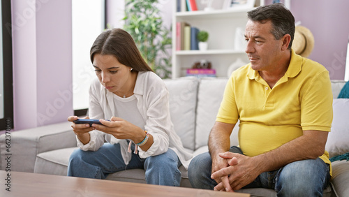 Huddled together at home, father and daughter share a serene indoor moment, sitting on the sofa, engrossed in using smartphone technology - a cherished family bond sustained even in the digital age.