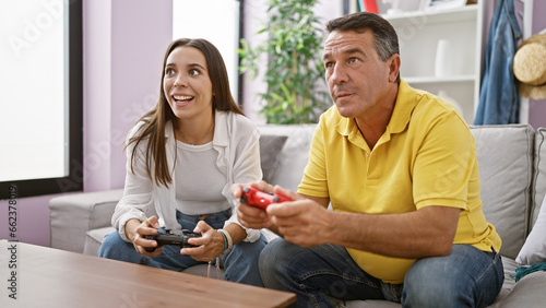 Hispanic father and daughter enjoy bonding time playing a video game at home, sitting on the living room sofa, smiles of joy on their faces, as they confidently use their gamepad controls.