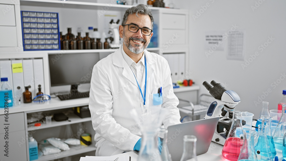 Attractive, grey-haired young hispanic man, a confident scientist, all smiles while conducting a biology analysis using his laptop in a bustling lab. Attractive, grey-haired young hispanic man, a confident scientist, all smiles while conducting a biology analysis using his laptop in a bustling lab.