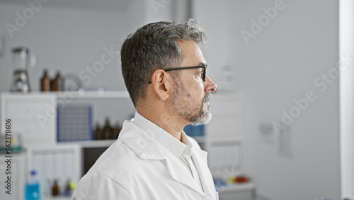 Unwavering young hispanic man  a grey-haired scientist  immersed in serious research  standing side-angled in the bustling laboratory.