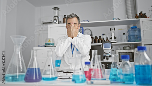 Stressed out grey-haired, young hispanic man, a professional scientist, under the microscope's spell, caught in overwork at an indoor laboratory