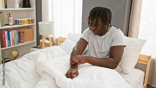 Bild auf Leinwand African american man scratching arm for itchy sitting on bed at bedroom