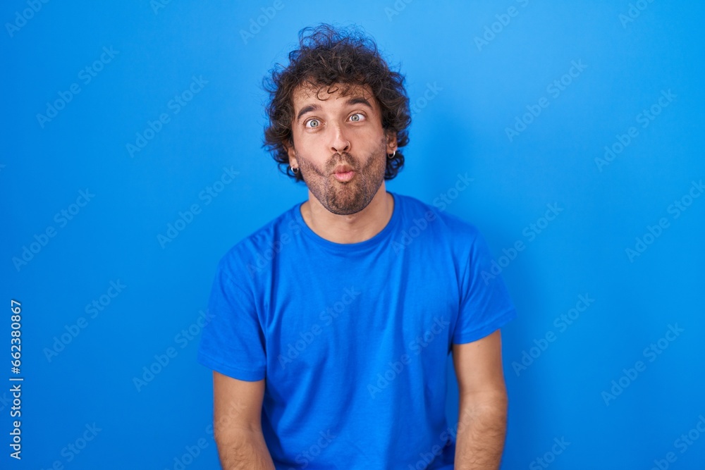 Hispanic young man standing over blue background making fish face with lips, crazy and comical gesture. funny expression. Hispanic young man standing over blue background making fish face with lips, crazy and comical gesture. funny expression.