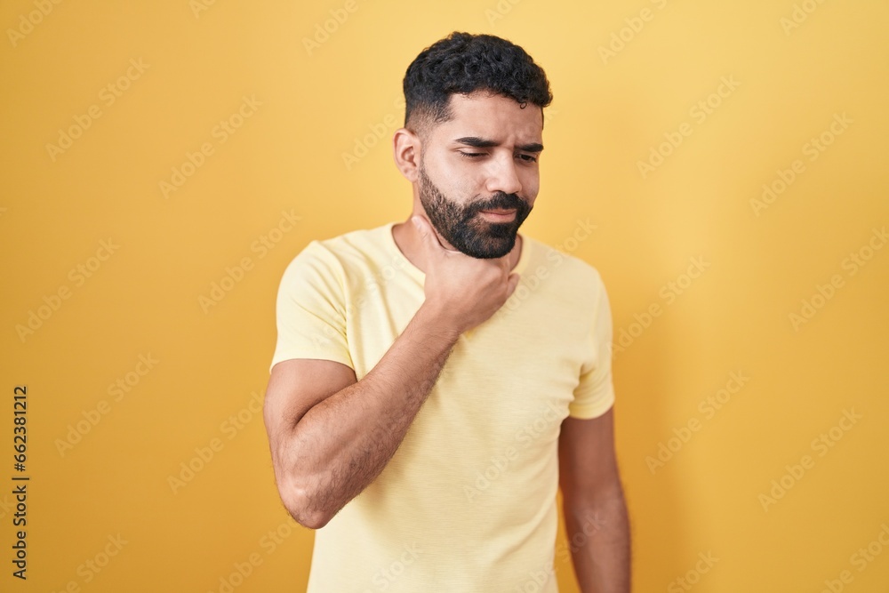Hispanic man with beard standing over yellow background touching painful neck, sore throat for flu, clod and infection Hispanic man with beard standing over yellow background touching painful neck, sore throat for flu, clod and infection