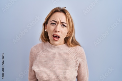 Hispanic woman standing over blue background in shock face, looking skeptical and sarcastic, surprised with open mouth