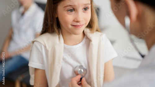 Doctor and kid patient are in the clinic. Physician in white coat examining a smiling young girl with a stethoscope, close up. Medicine, therapy concept