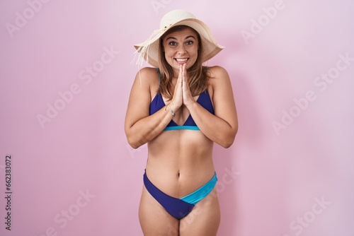 Young hispanic woman wearing bikini over pink background praying with hands together asking for forgiveness smiling confident.
