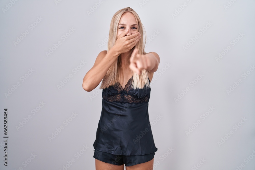Young caucasian woman wearing nightgown laughing at you, pointing finger to the camera with hand over mouth, shame expression Young caucasian woman wearing nightgown laughing at you, pointing finger to the camera with hand over mouth, shame expression