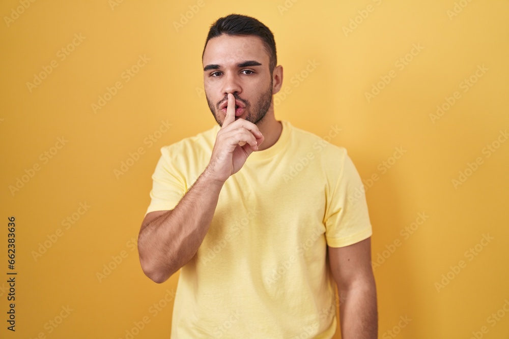 Young hispanic man standing over yellow background asking to be quiet with finger on lips. silence and secret concept. Young hispanic man standing over yellow background asking to be quiet with finger on lips. silence and secret concept.