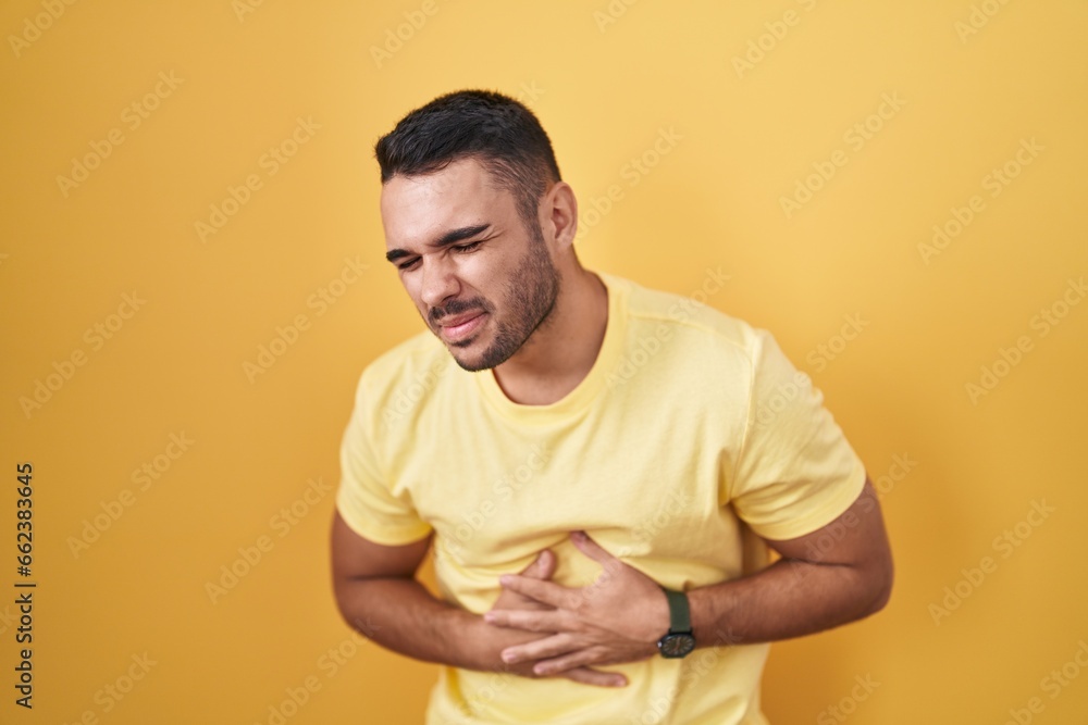 Young hispanic man standing over yellow background with hand on stomach because indigestion, painful illness feeling unwell. ache concept. Young hispanic man standing over yellow background with hand on stomach because indigestion, painful illness feeling unwell. ache concept.