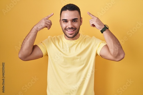 Young hispanic man standing over yellow background smiling pointing to head with both hands finger, great idea or thought, good memory