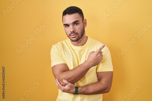 Young hispanic man standing over yellow background pointing with hand finger to the side showing advertisement, serious and calm face