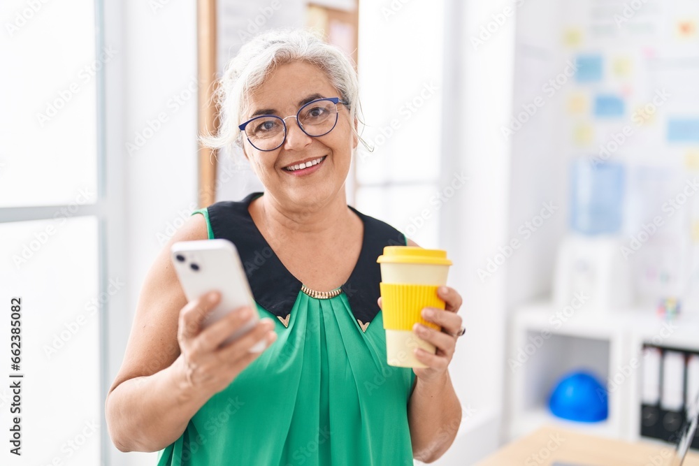 Middle age grey-haired woman business worker using smartphone drinking coffee at office Middle age grey-haired woman business worker using smartphone drinking coffee at office