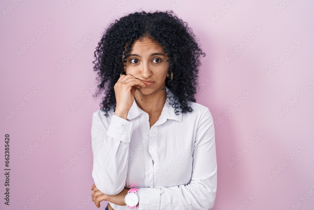 Hispanic woman with curly hair standing over pink background looking stressed and nervous with hands on mouth biting nails. anxiety problem. Hispanic woman with curly hair standing over pink background looking stressed and nervous with hands on mouth biting nails. anxiety problem.