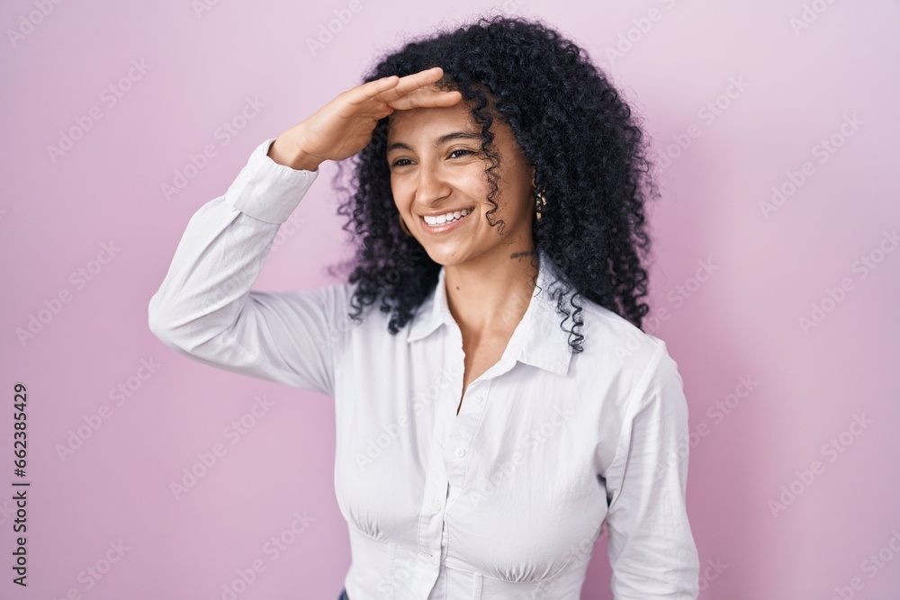 Hispanic woman with curly hair standing over pink background very happy and smiling looking far away with hand over head. searching concept. Hispanic woman with curly hair standing over pink background very happy and smiling looking far away with hand over head. searching concept.