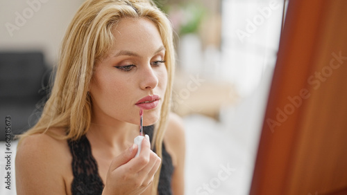 Young blonde woman putting lipstick sitting on bed looking sexy at mirror at bedroom