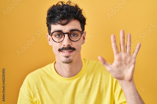 Hispanic man wearing glasses standing over yellow background showing and pointing up with fingers number five while smiling confident and happy.