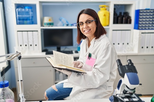 Young beautiful hispanic woman scientist smiling confident reading book at laboratory