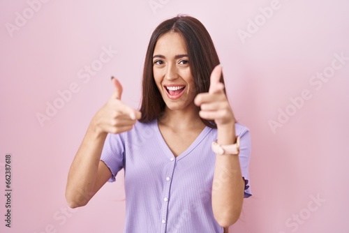 Young hispanic woman with long hair standing over pink background pointing fingers to camera with happy and funny face. good energy and vibes.