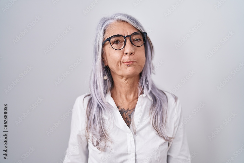 Middle age woman with tattoos wearing glasses standing over white background puffing cheeks with funny face. mouth inflated with air, crazy expression.