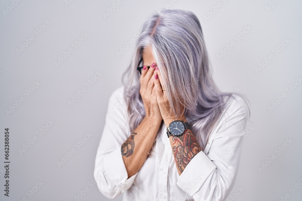 Middle age woman with tattoos wearing glasses standing over white background with sad expression covering face with hands while crying. depression concept. Middle age woman with tattoos wearing glasses standing over white background with sad expression covering face with hands while crying. depression concept.
