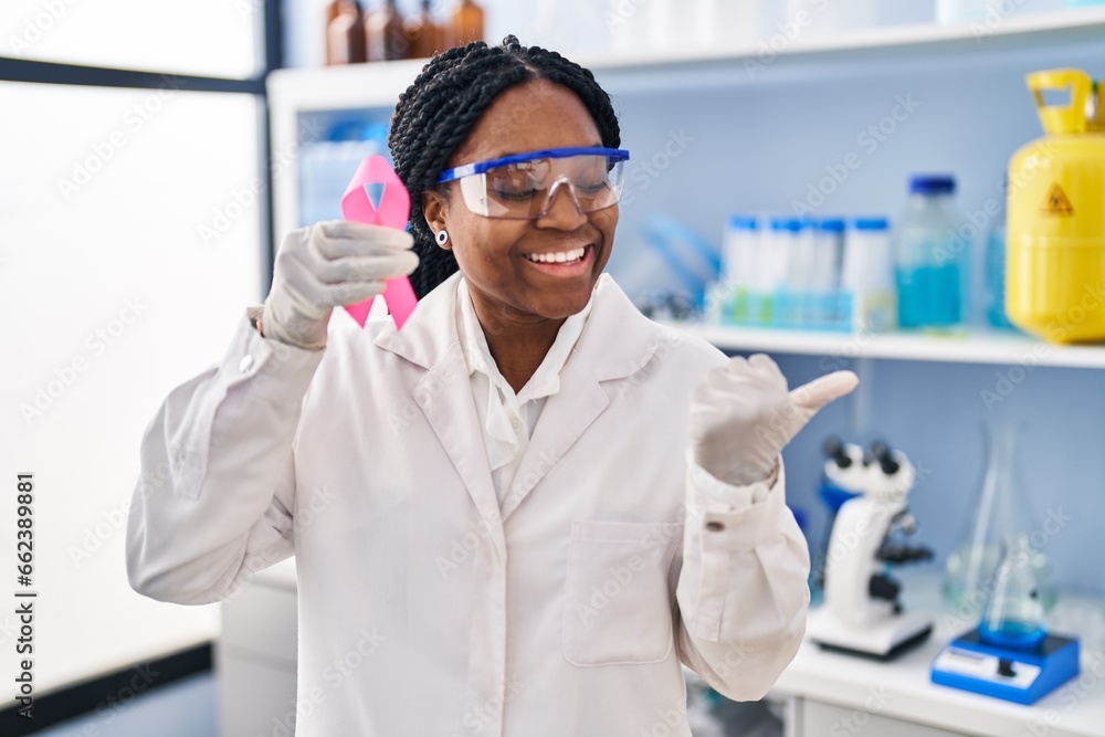 Naklejka premium African american woman working at scientist laboratory holding pink ribbon pointing thumb up to the side smiling happy with open mouth
