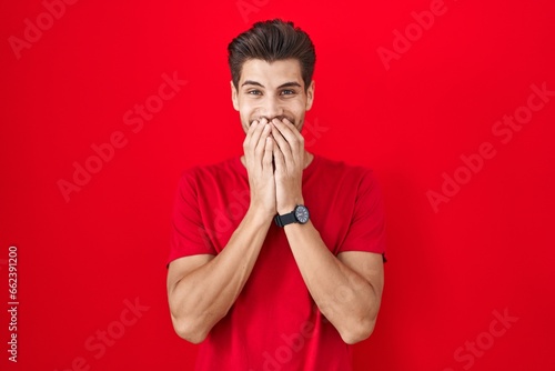 Fotografie Young hispanic man standing over red background laughing and embarrassed giggle