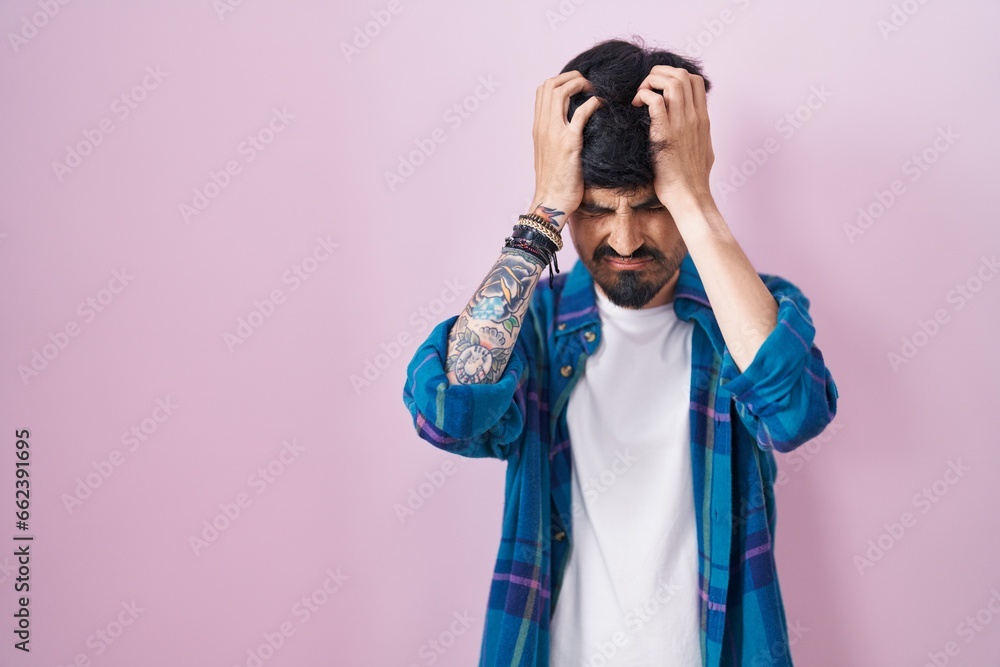 Young hispanic man with beard standing over pink background suffering from headache desperate and stressed because pain and migraine. hands on head. Young hispanic man with beard standing over pink background suffering from headache desperate and stressed because pain and migraine. hands on head.