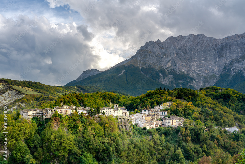 Quaint hillside village of Castelli in Abruzzo, nestled against majestic mountains. Sunlight bathes the historic homes surrounded by lush greenery under a dramatic sky.