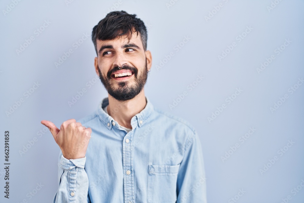 Young hispanic man with beard standing over blue background smiling with happy face looking and pointing to the side with thumb up. Young hispanic man with beard standing over blue background smiling with happy face looking and pointing to the side with thumb up.
