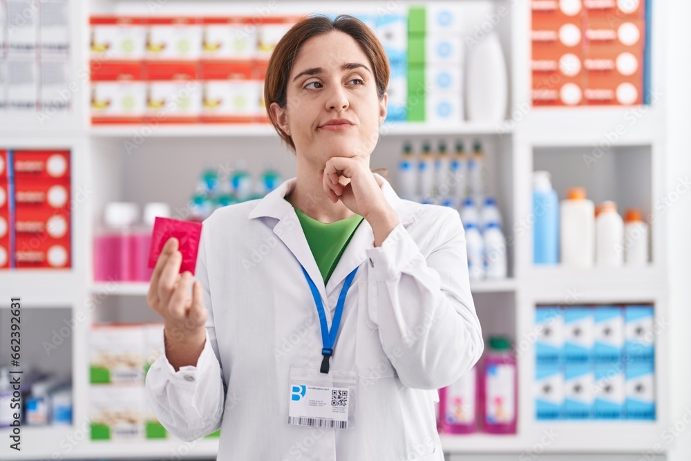 Brunette woman working at pharmacy drugstore holding condom serious face thinking about question with hand on chin, thoughtful about confusing idea