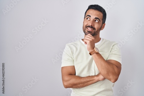 Hispanic man with beard standing over isolated background with hand on chin thinking about question, pensive expression. smiling and thoughtful face. doubt concept.