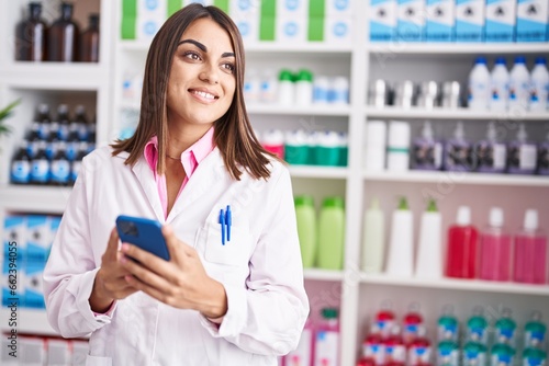 Young beautiful hispanic woman pharmacist using smartphone working at pharmacy