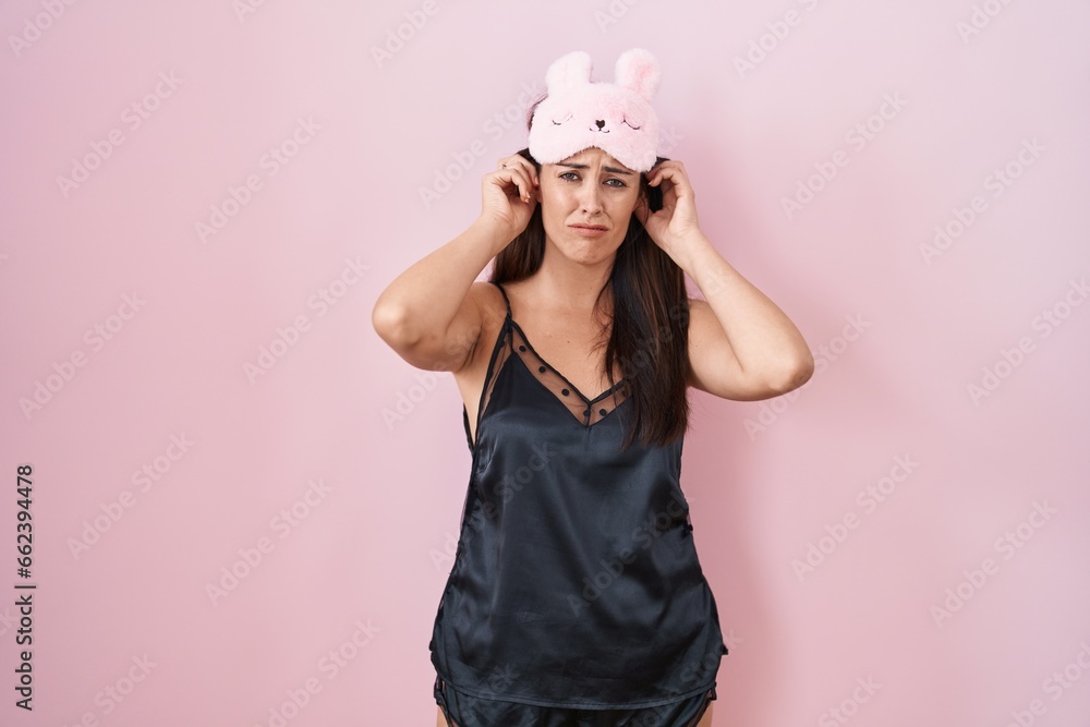 Young brunette woman wearing sleep mask and pyjama covering ears with fingers with annoyed expression for the noise of loud music. deaf concept. Young brunette woman wearing sleep mask and pyjama covering ears with fingers with annoyed expression for the noise of loud music. deaf concept.