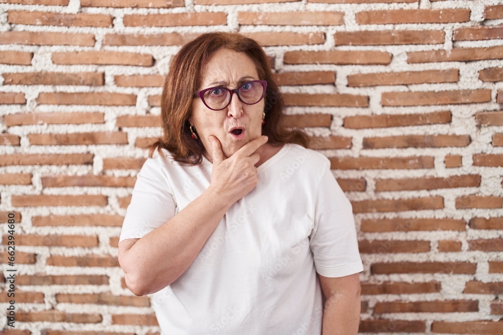 Senior woman with glasses standing over bricks wall looking fascinated with disbelief, surprise and amazed expression with hands on chin Senior woman with glasses standing over bricks wall looking fascinated with disbelief, surprise and amazed expression with hands on chin