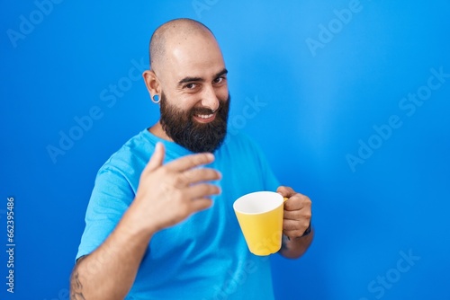 Young hispanic man with beard and tattoos drinking a cup of coffee beckoning come here gesture with hand inviting welcoming happy and smiling © Krakenimages.com