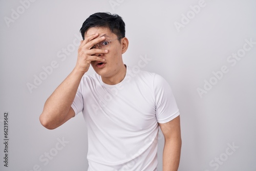 Young asian man standing over white background peeking in shock covering face and eyes with hand, looking through fingers with embarrassed expression.