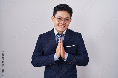 Young asian man wearing business suit and tie praying with hands together asking for forgiveness smiling confident.