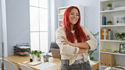 Smiling young redhead woman confidently standing with arms crossed at office, an embodiment of business success.
