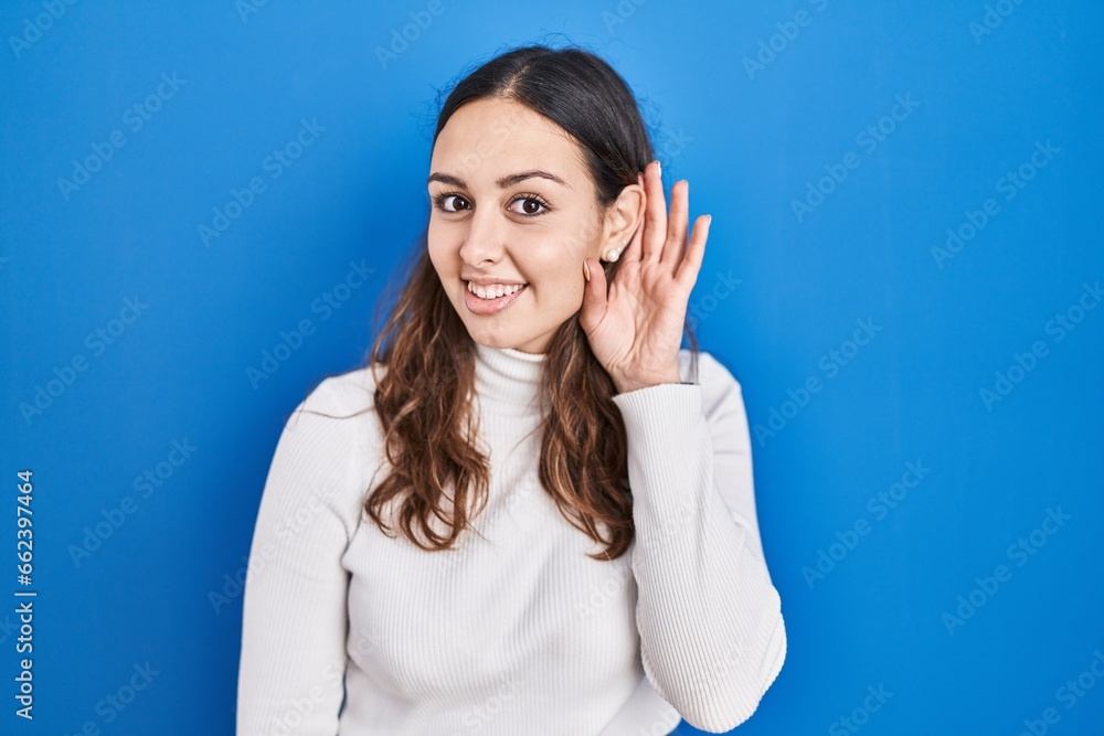 Young hispanic woman standing over blue background smiling with hand over ear listening an hearing to rumor or gossip. deafness concept. Young hispanic woman standing over blue background smiling with hand over ear listening an hearing to rumor or gossip. deafness concept.