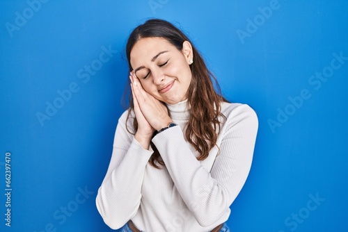 Young hispanic woman standing over blue background sleeping tired dreaming and posing with hands together while smiling with closed eyes.