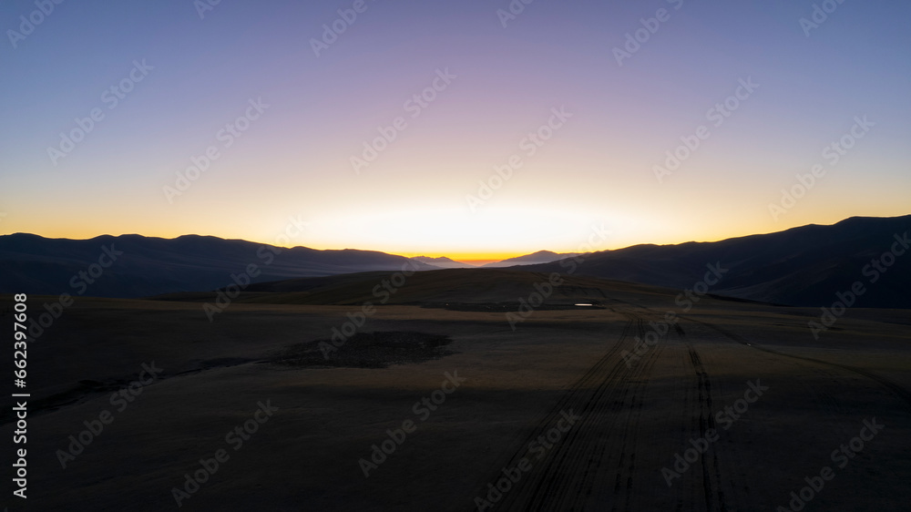 Dawn in the mountains. The violet-orange color of the sky. The red rays of the sun come out from behind the peaks. A light haze in the gorge. Traces of the road and puddles are visible. Drone view