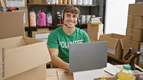 Handsome young hispanic man volunteering at a community charity center, cheerfully smiling while sitting at a table, using laptop with headphones
