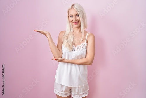 Caucasian woman wearing pajama wearing pink background smiling cheerful presenting and pointing with palm of hand looking at the camera.